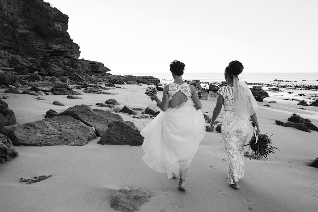 178 - Two brides walking down the beach holding hands on their wedding day.