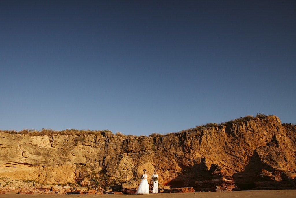 179 - Two brides in white dresses standing barefoot in the sand in front of red cliff at Eco Beach in Broome, Western Australia.