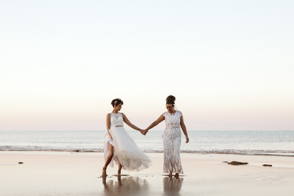 182 - Two brides holding hand on the beach on their wedding day in white brides dresses.
