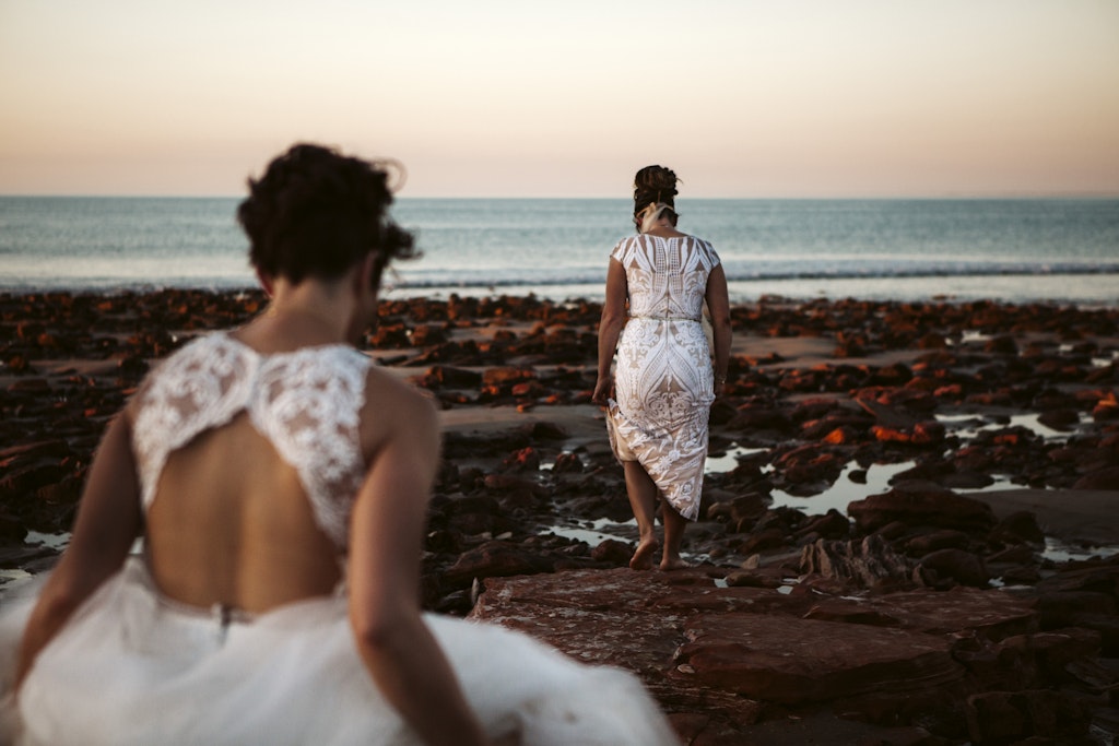 183 - Brides walking on the beach with no shoes in their wedding dresses at Eco Beach.