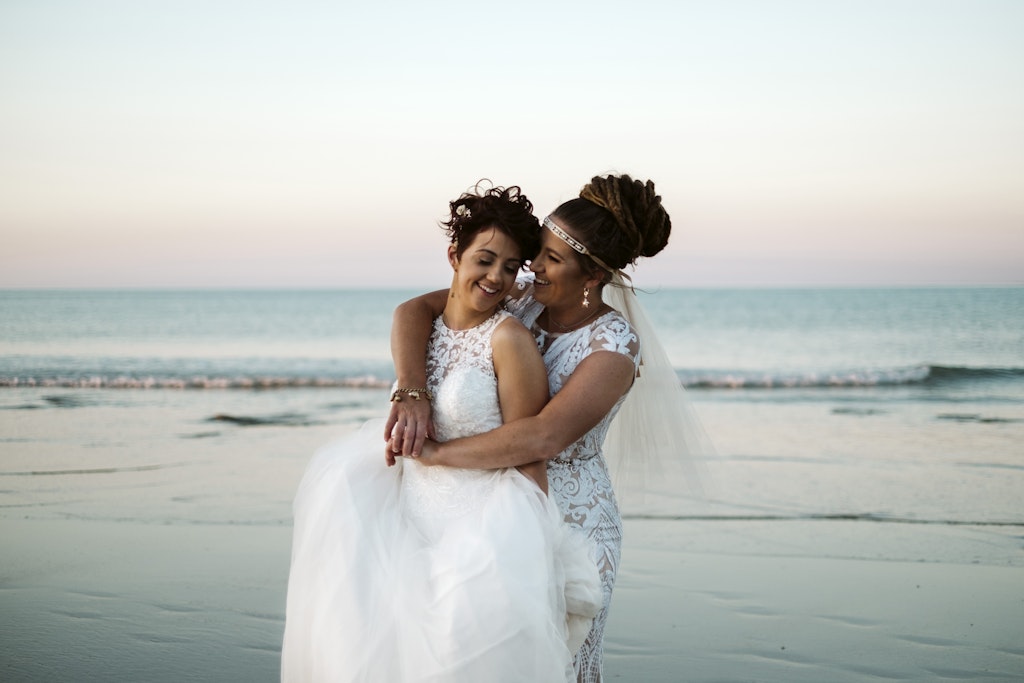 187 - Two brides in white wedding dresses on their wedding day standing on a beach with the ocean in the background.