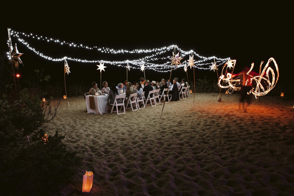 198 - wedding reception dinner on beach with long table and fairy lighting and man fire twirling on the side at eco beach in Broome western australia.