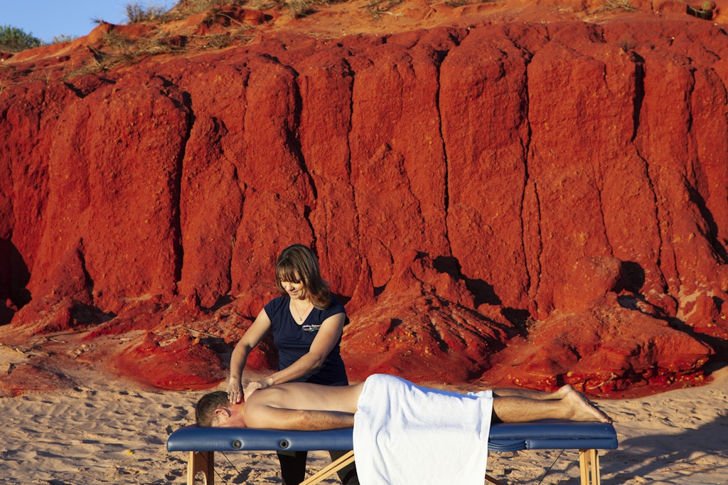 63 - female massage therapist massaging back of man on beach with red cliff in the back ground. Broome, Western Australia