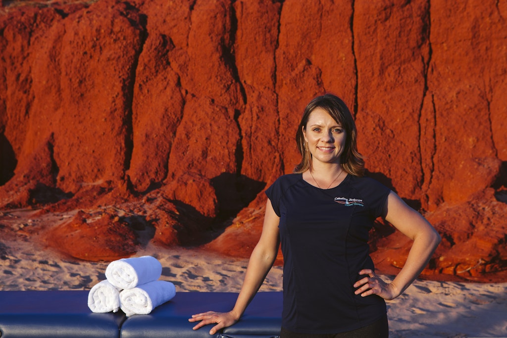 71 - Young female massage therapist with short dark hair standing with massage table in front of red cliffs on the beach.