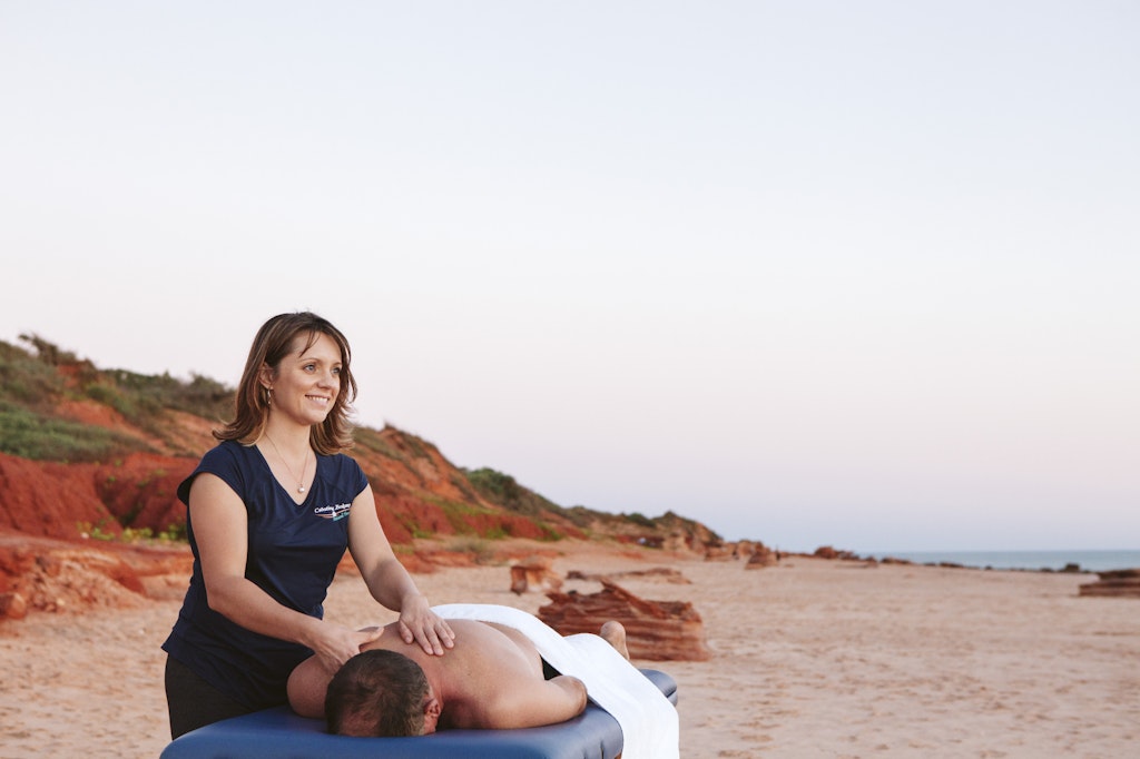 88 - Young female massage therapist massaging male on table on white sand looking out into the ocean.