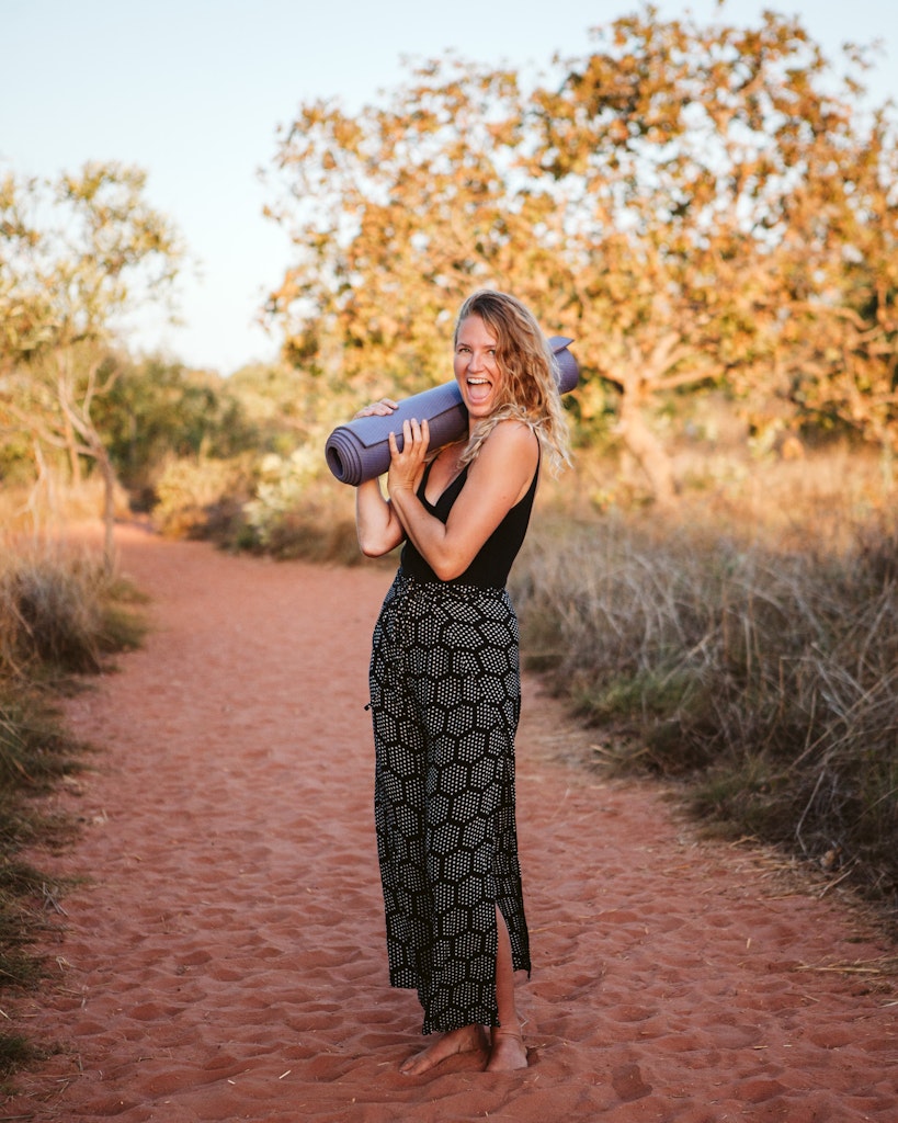08 - Girl with long blonde hair holding yoga mat with big smile looking happy standing on red sand path in Broome.