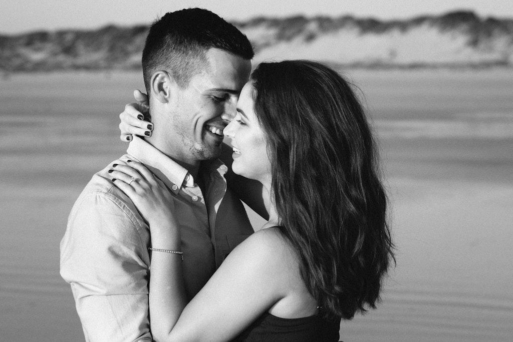 11 - Black and white image of newly engaged couple cuddling on beach showing of engagement ring.