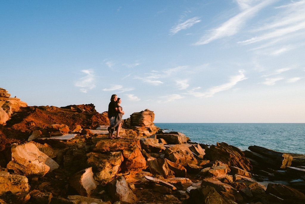 17 - Newly engaged couple standing on red rocks over looking ocean in Broome, Western Australia.